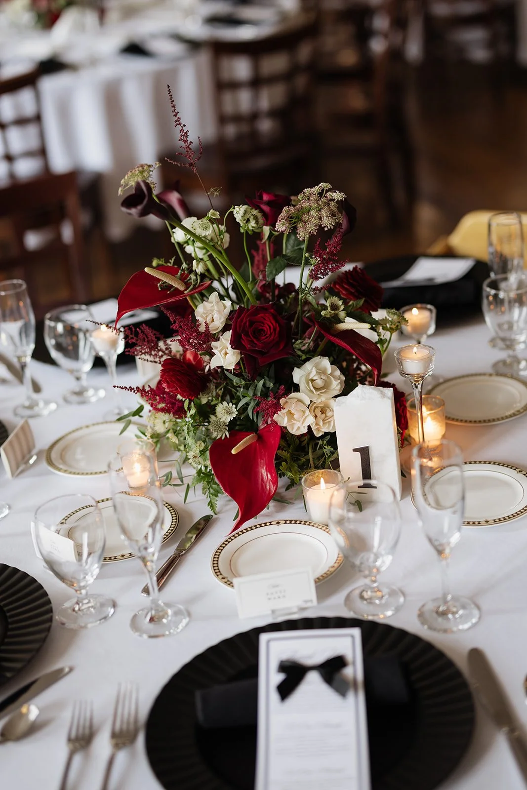 A table decorated with a floral centerpiece of red and white flowers, surrounded by lit candles, and set with elegant plates, glasses, and cutlery at a formal event or wedding reception.