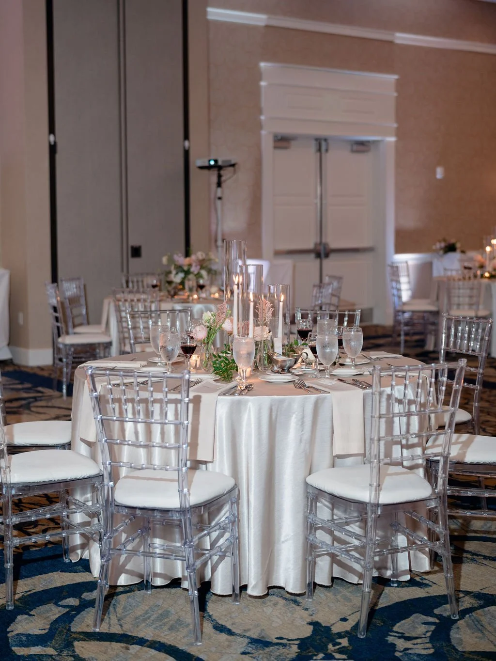 Elegant banquet table set with white tablecloth, floral centerpieces, tall candles, wine and water glasses, and silverware in a decorated event hall.