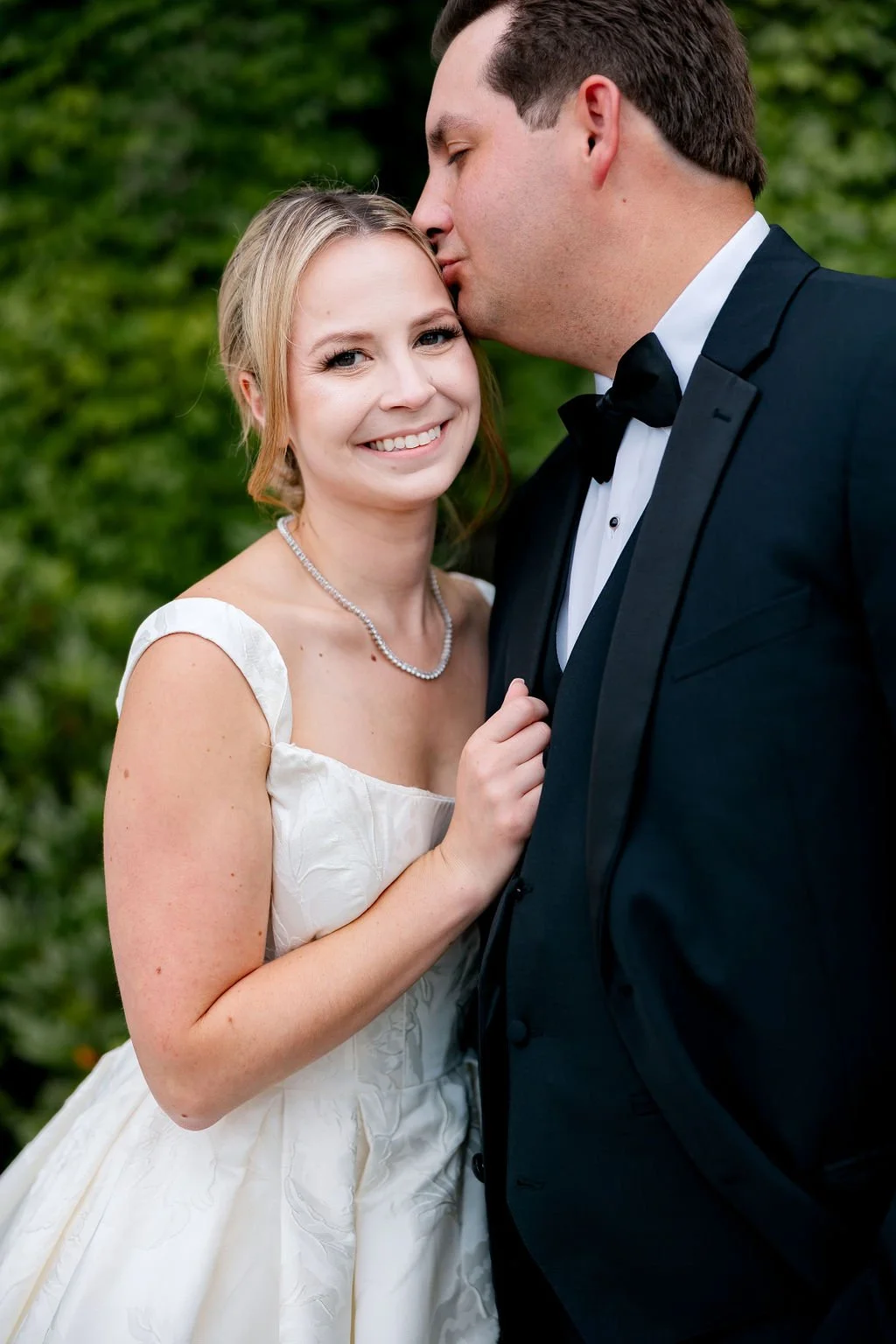 A newlywed couple in wedding attire, the woman smiling at the camera and the man kissing her forehead, outdoors with green foliage in the background.