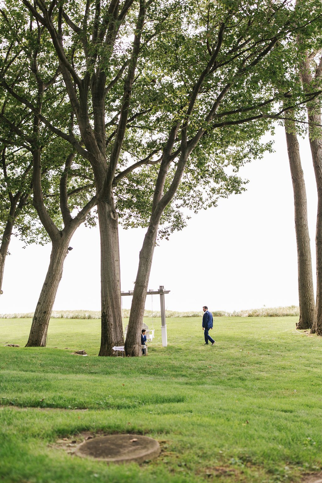A man in a blue suit walking on a green lawn near trees under a cloudy sky.