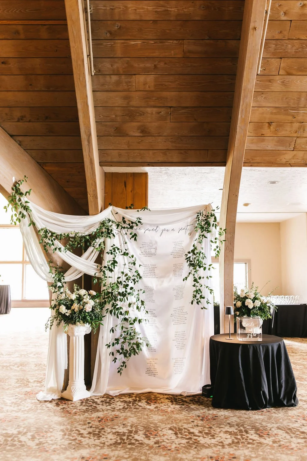 A wedding seating chart display, with a white fabric backdrop with black calligraphy, greenery accents, and floral arrangements on white pillars, inside a rustic venue with wooden ceiling and natural light.