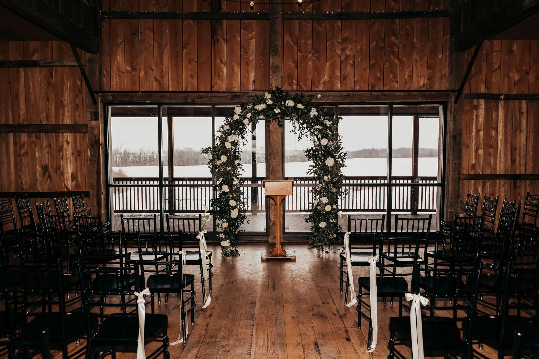 Interior of a rustic wedding venue with wooden walls, black chairs arranged in rows, and a floral arch at the front near large windows overlooking a snowy landscape.