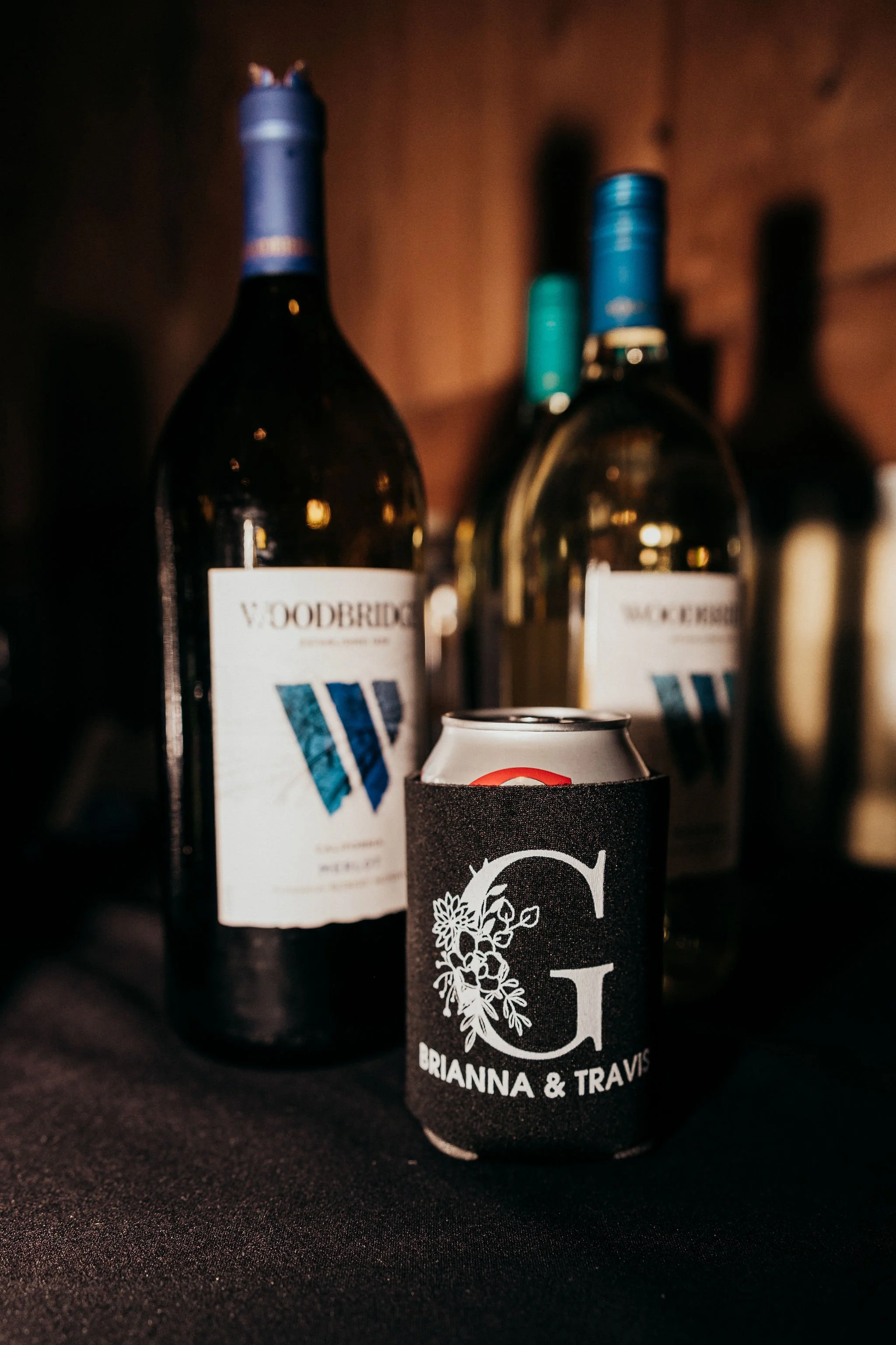 Three bottles of wine with dark labels and colorful foil tops, and a black drink koozie with white floral design and text, all set against a dark background.