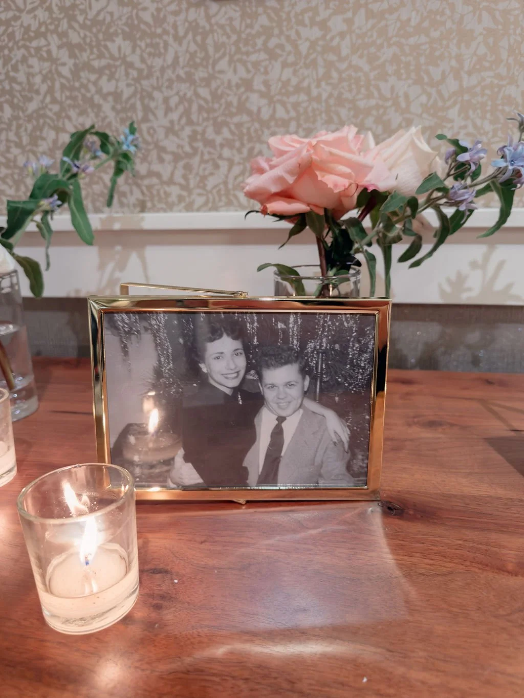 A framed black and white photograph of a man and a woman, along with a pink rose and purple flowers in a glass vase, and a lit candle on a wooden surface.