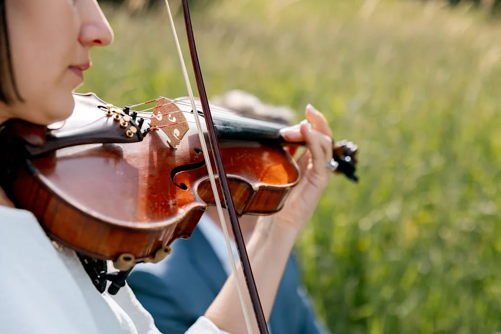 A person playing a violin outdoors with grassy background.