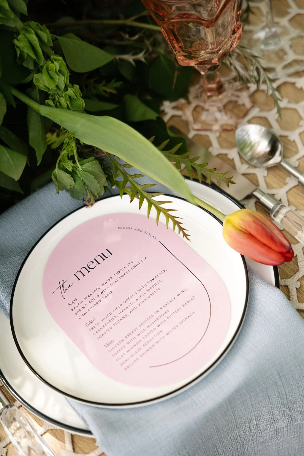A table setting featuring a pink menu on a white plate, decorated with assorted green foliage and a single orange-pink tulip, with a spoon, a small glass, and a patterned tablecloth.