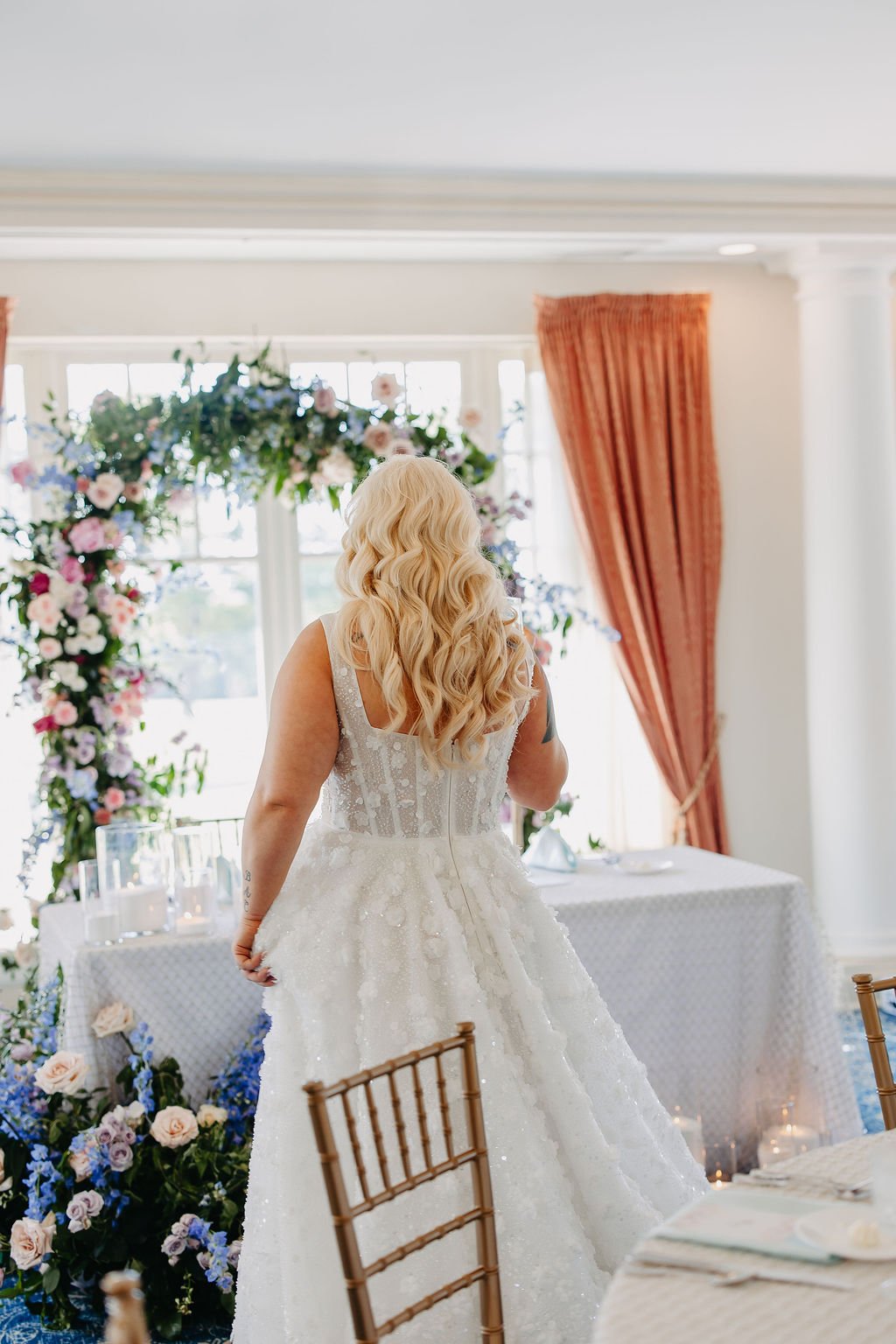 A woman with long blonde hair wearing a wedding dress stands in a decorated room with floral arrangements and large windows, possibly preparing for a wedding.