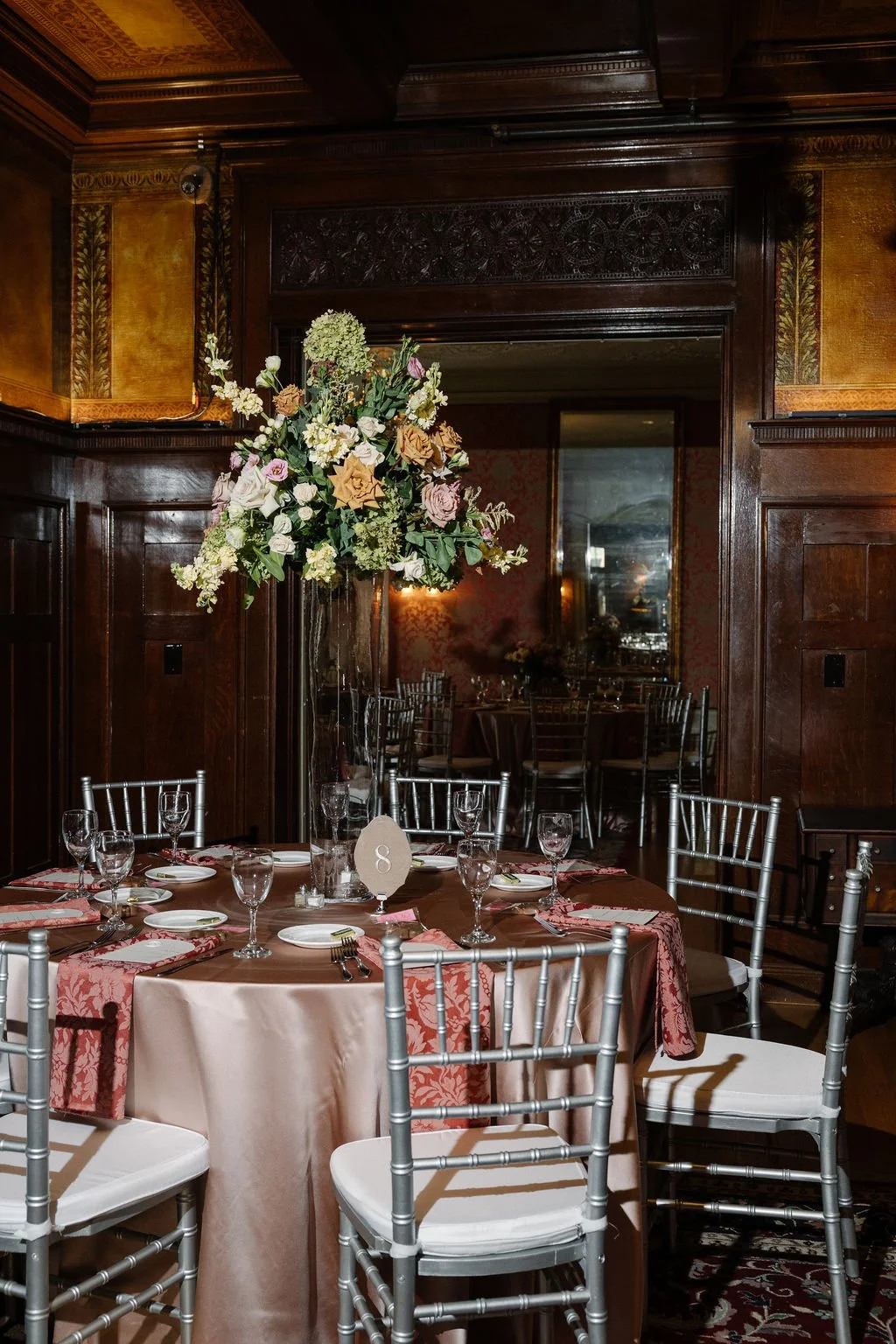 A formal dining room with a round table decorated with a large floral centerpiece, set with plates, silverware, and glassware, surrounded by silver Chiavari chairs, in a wood-paneled, ornately decorated room with gold accents.