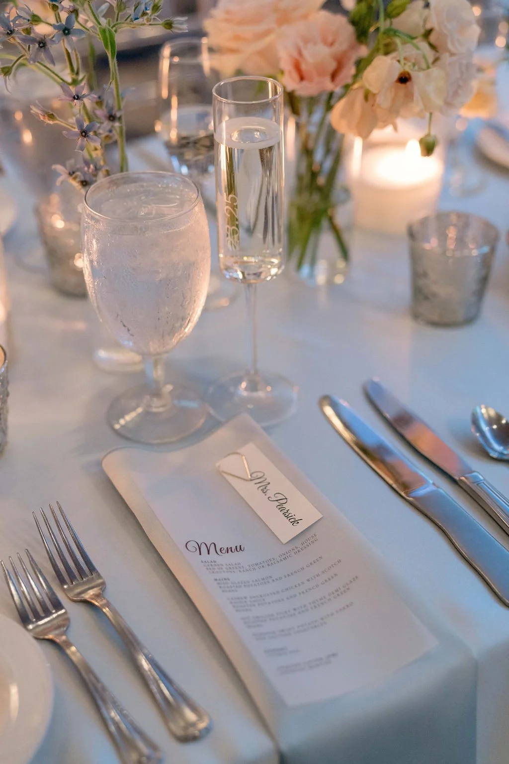 Elegant table setting at a wedding reception with glasses of water and champagne, floral arrangements, candles, a menu, and silverware.