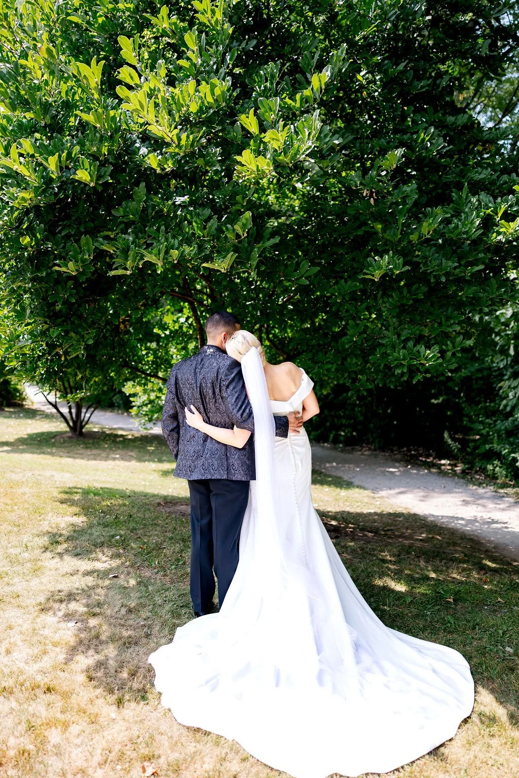 A bride and groom embracing under a large green tree on their wedding day.