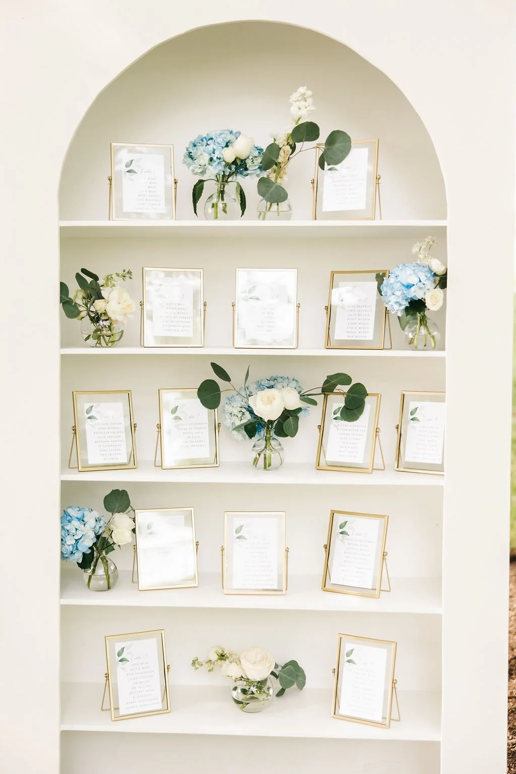 A white display shelf with five levels, decorated with glass vases holding white and blue flowers and framed documents with gold frames, some with green leaf illustrations.