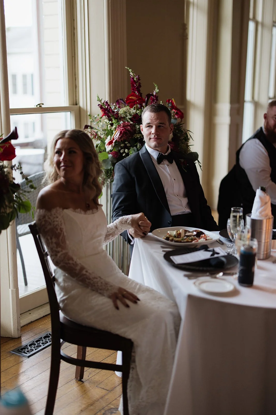 A bride and groom holding hands at a formal wedding reception, sitting at a table with plates of food, in a room with large windows and floral decorations.