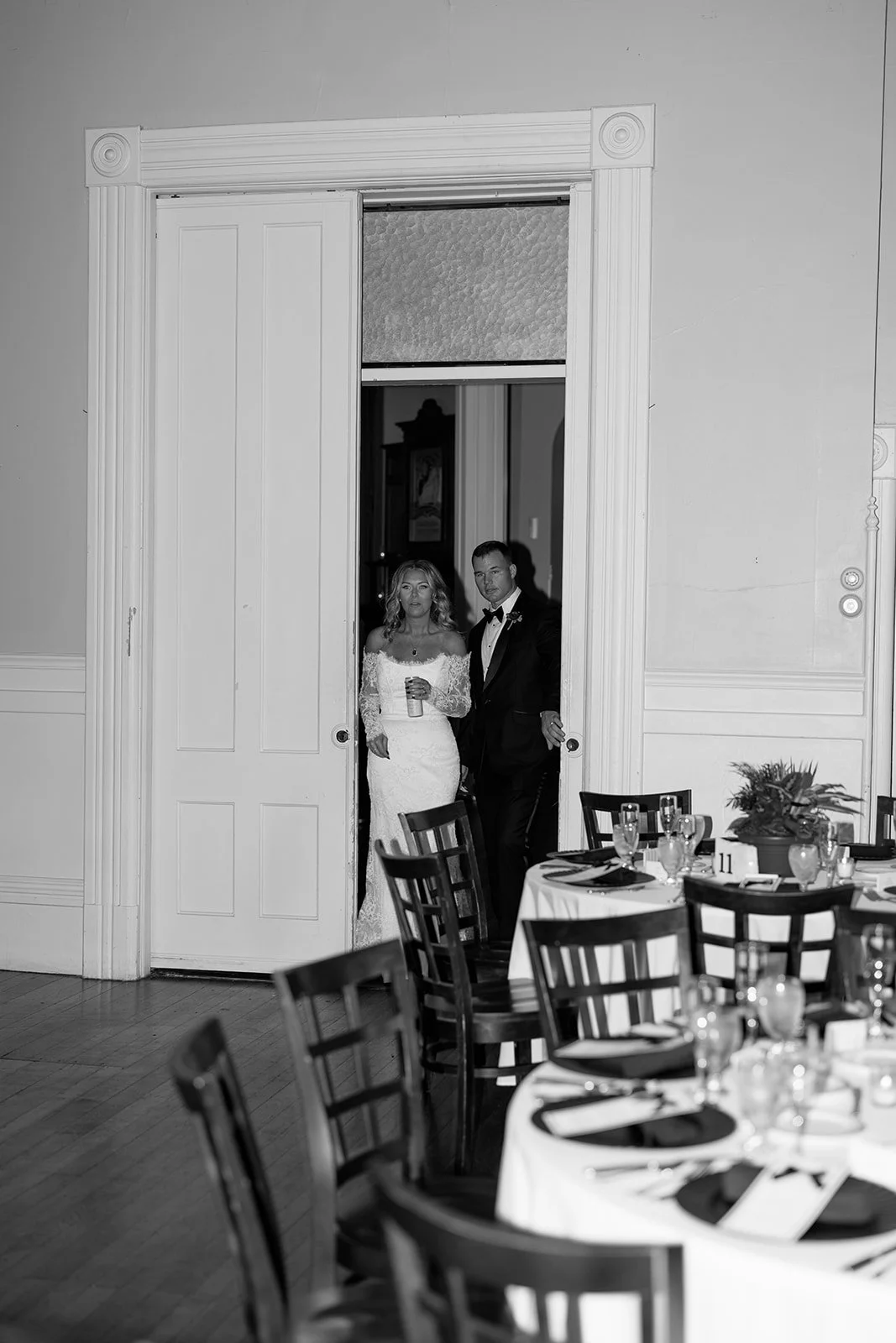A black and white photo of a bride in a wedding gown and a groom in a tuxedo standing at a doorway during a wedding reception.