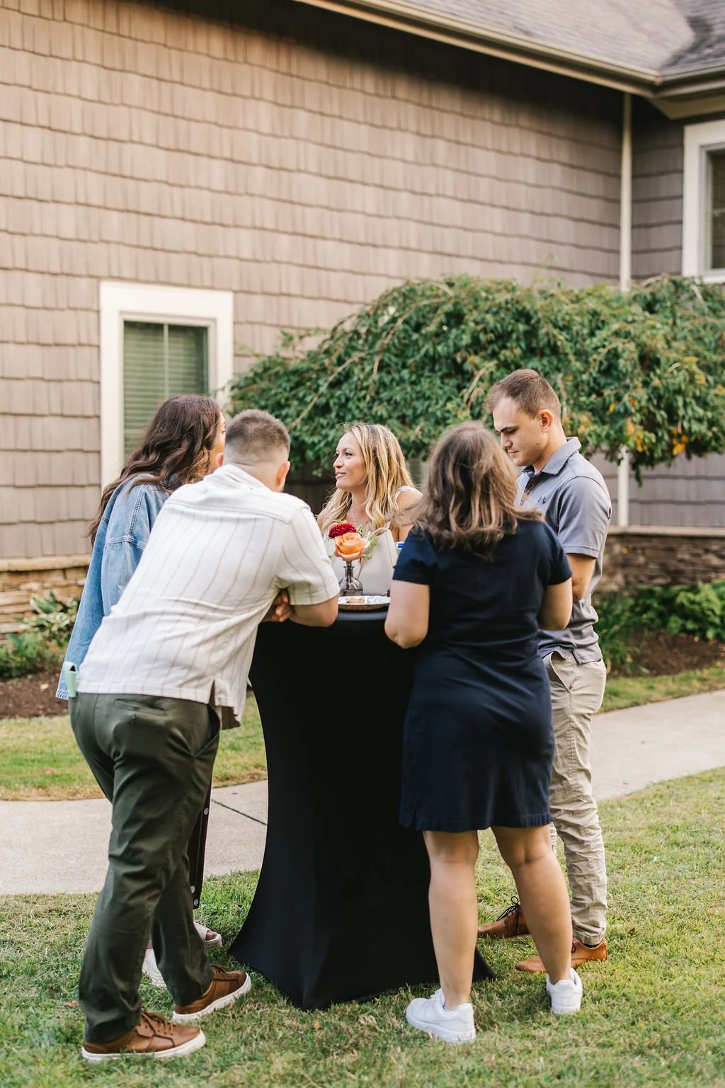 Group of five young adults gathered outdoors around a small table with a centerpiece, engaged in conversation, in front of a house with a landscaped yard.