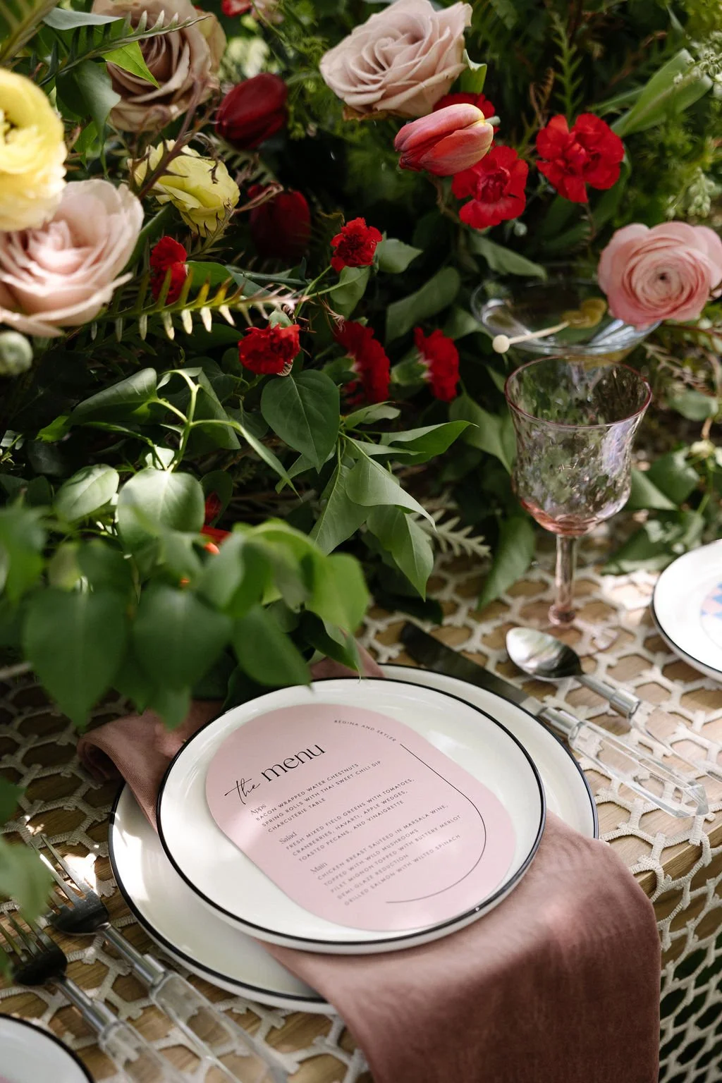 A table setting with a floral centerpiece featuring pink roses, red carnations, and green foliage. There are plates, a pink napkin, silverware, and a pink-tinted glass on the table. The plate has a menu with elegant typography.