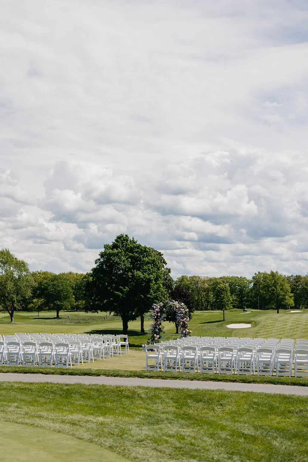 Outdoor wedding setup with white chairs arranged on a grassy field, decorated arch with flowers, trees, and sand bunkers in the background under a cloudy sky.