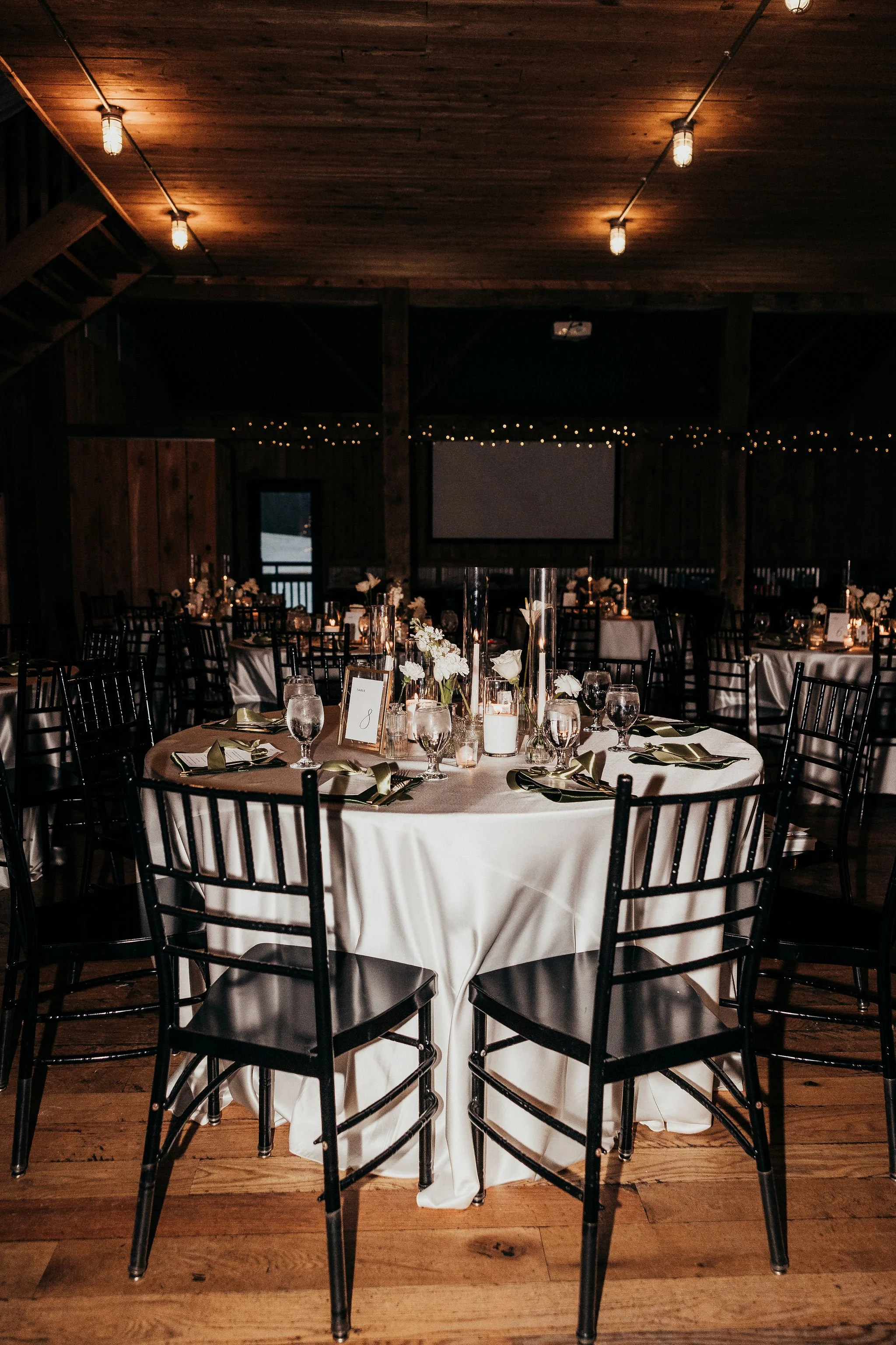 Event space decorated for a wedding reception with round tables, white tablecloths, floral centerpieces, and black chairs, in a rustic wooden venue with warm lighting and string lights.