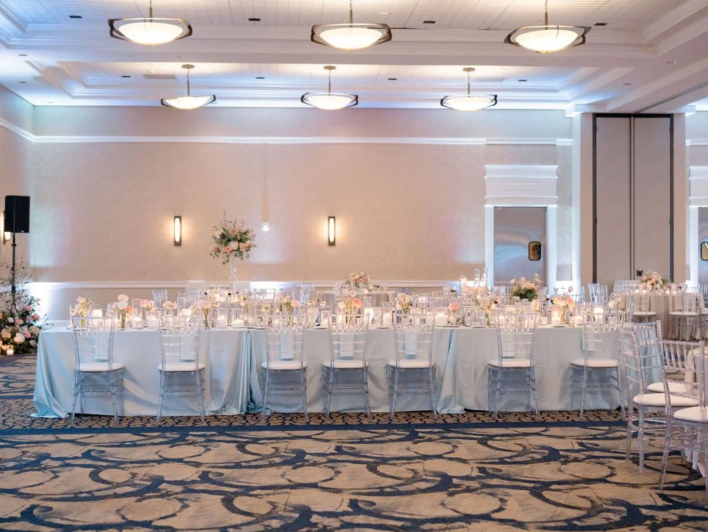 Elegant banquet hall with round tables covered in white tablecloths, decorated with floral centerpieces, set with glassware and candles, surrounded by transparent chairs, and illuminated by soft lighting and ceiling fixtures.
