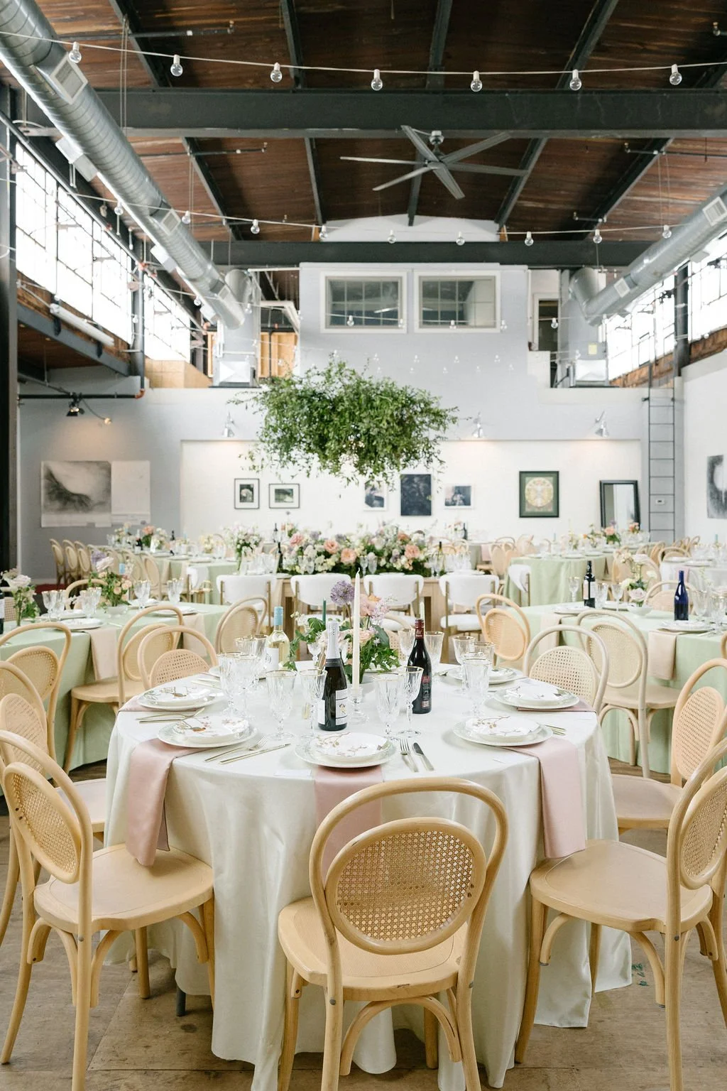 Elegant event space with round tables draped in white cloths, adorned with floral centerpieces, wine bottles, and place settings, under string lights in a modern industrial-style venue.