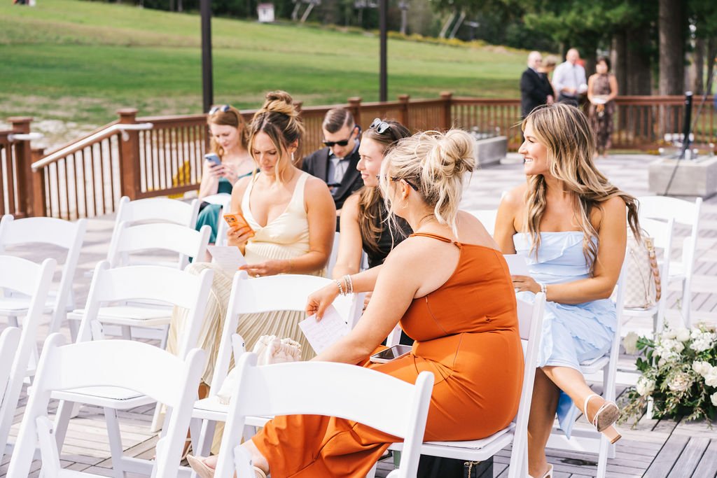 People sitting on white chairs outdoors, possibly waiting for an event, with a grassy area and trees in the background.