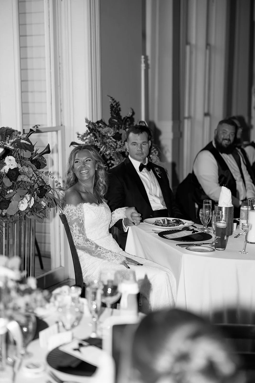 Black and white photo of a bride and groom sitting at a wedding reception table, holding hands. The bride is smiling widely, wearing a lace wedding dress, and the groom has a serious expression, dressed in a tuxedo with a bow tie. There are floral ar
