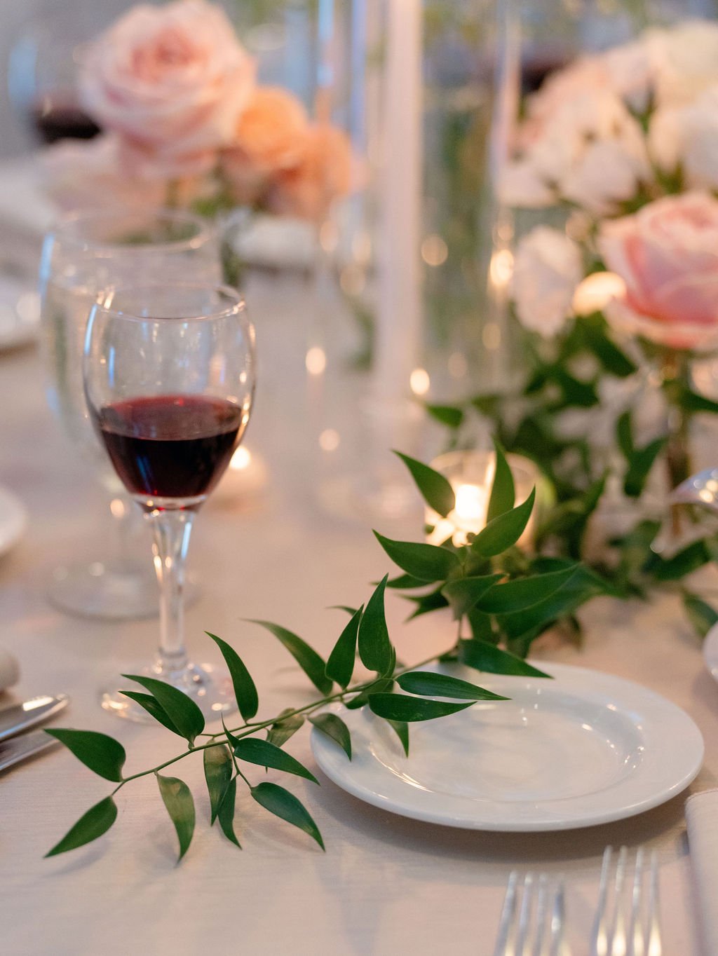 A close-up of a elegantly set dining table with a glass of red wine, decorative greenery, and floral arrangements with pink roses in the background.