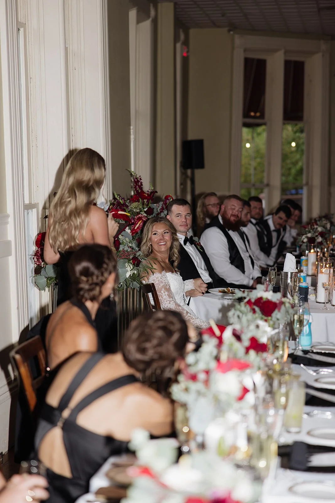 At a wedding reception, a woman is giving a toast while others listen, seated at a decorated table with floral centerpieces.