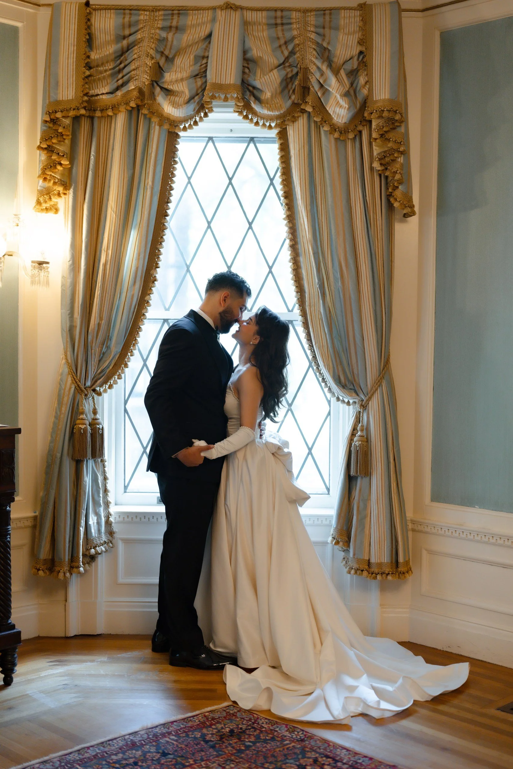 A bride and groom standing close together in front of a large window with ornate curtains, sharing a tender moment, in an elegant room with wood floors and decorative molding.
