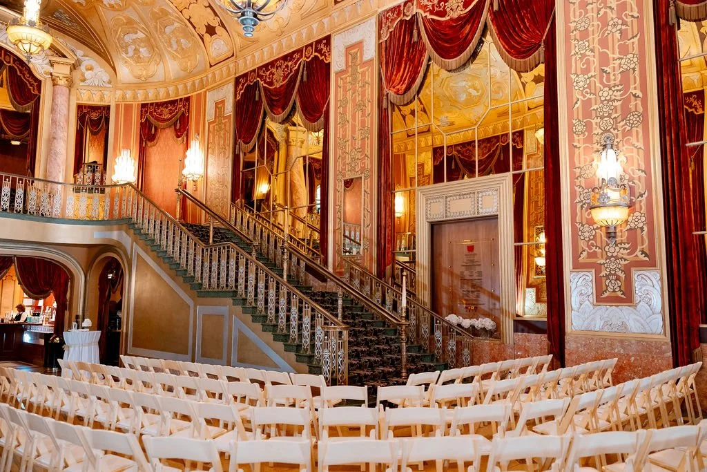 Elegant theater or ballroom with ornate gold and red decor, a curved staircase with intricate railings, chandeliers, and rows of empty white chairs.
