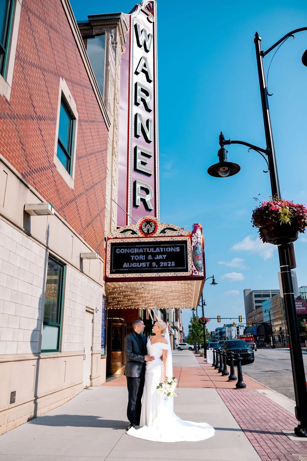 Bride and groom standing closely outside a theater marquee, with a sign congratulating Tori and Jay on August 8, 2019, under a clear blue sky.