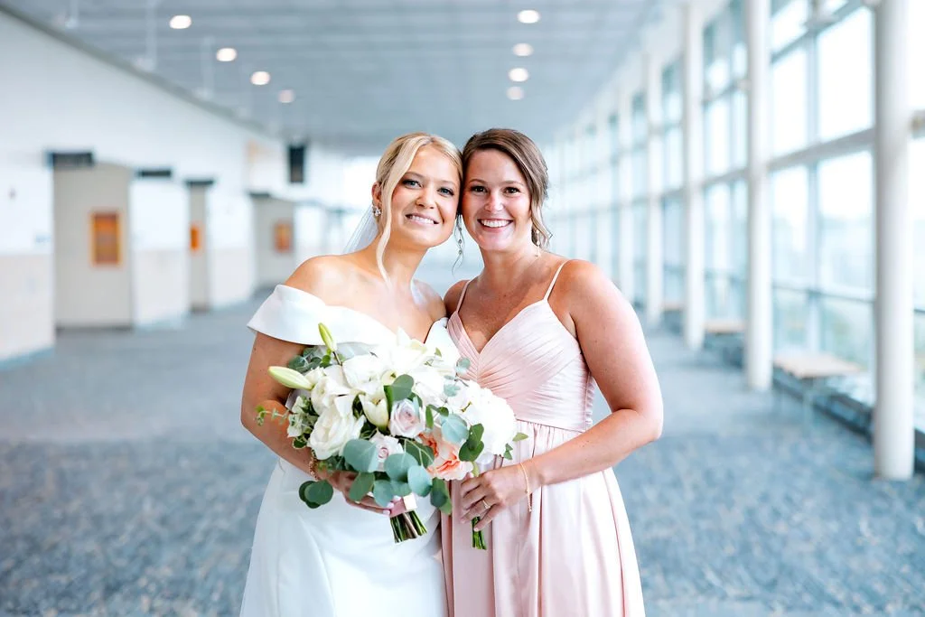 Two women smiling and posing together in a modern indoor setting with large windows, one wearing a white wedding dress holding a bouquet of white and pink flowers, and the other in a pale pink dress.