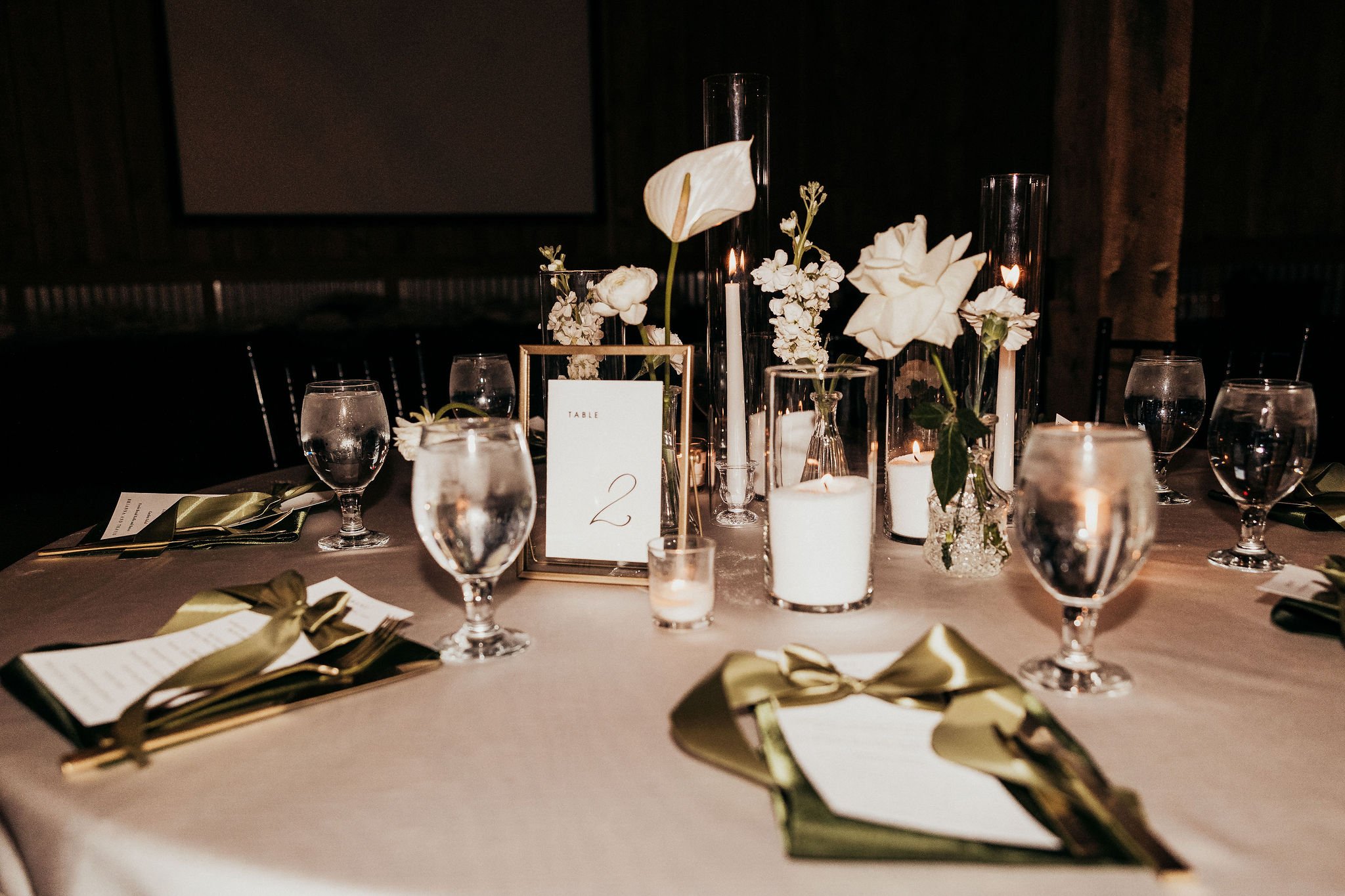 Elegant dinner table decorated with white flowers in glass vases, tall white candles, wine glasses filled with water, and table number card, set in a dimly lit room.
