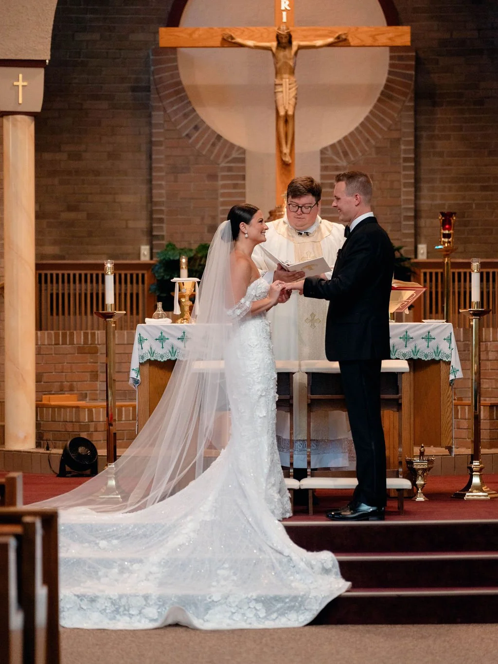 Wedding ceremony inside a church with a bride and groom exchanging vows at the altar, priest officiating, and a large crucifix with Jesus Christ in the background.