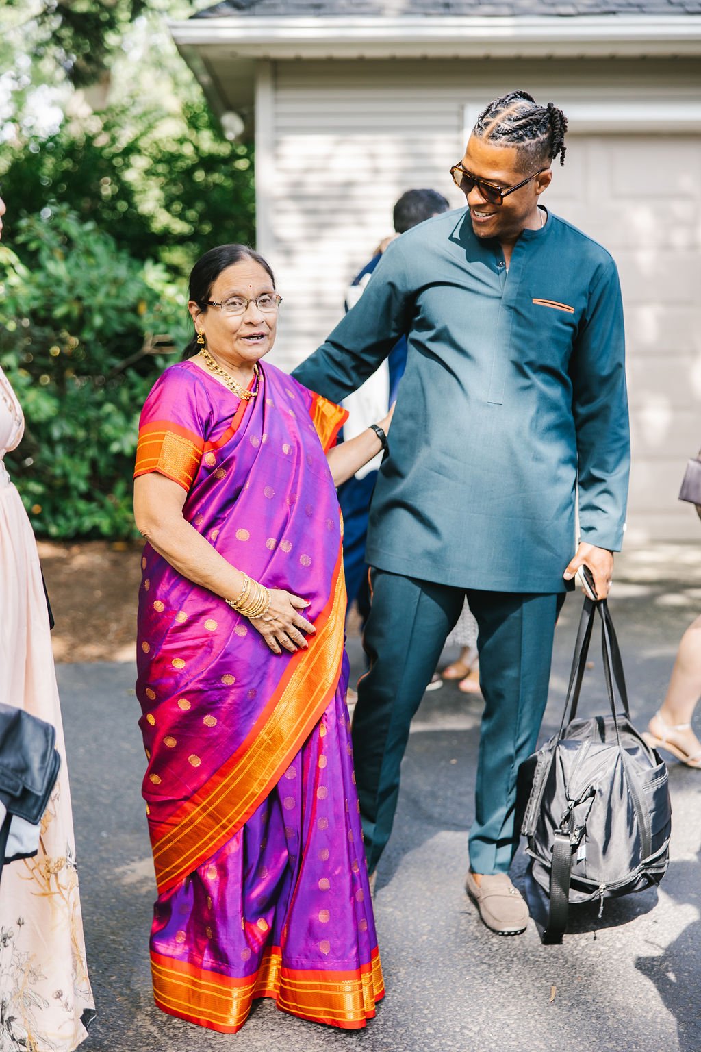 A woman in a purple and orange traditional sari talking to a smiling man in a blue suit with sunglasses, carrying a backpack, outdoors with greenery in the background.