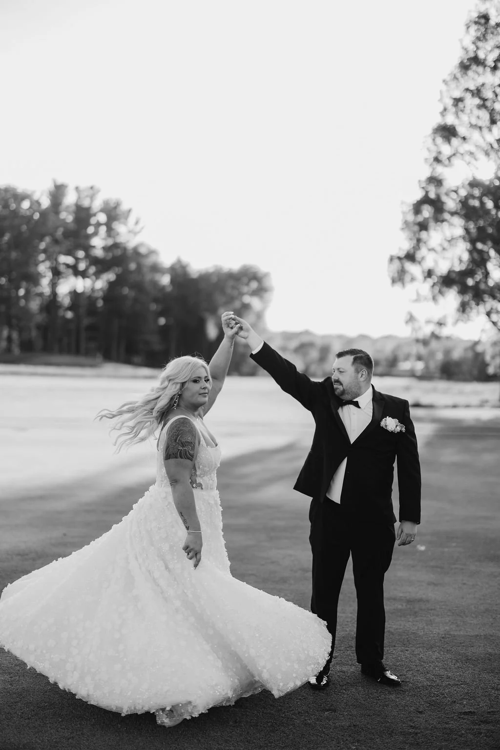 A black-and-white photo of a bride and groom dancing outdoors on a grassy field, with trees in the background. The groom is in a tuxedo, and the bride is in a wedding dress with tattoos visible on her arm. The groom lifts the bride's hand as they dan