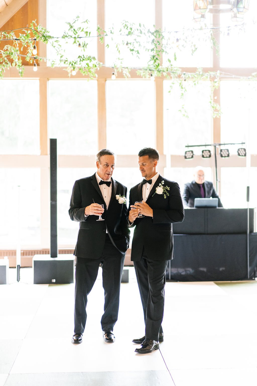 Two men in black tuxedos and bow ties, each with a white boutonniere, are standing together holding drinks at a wedding reception. They are indoors in a brightly lit space with large windows and hanging green foliage.