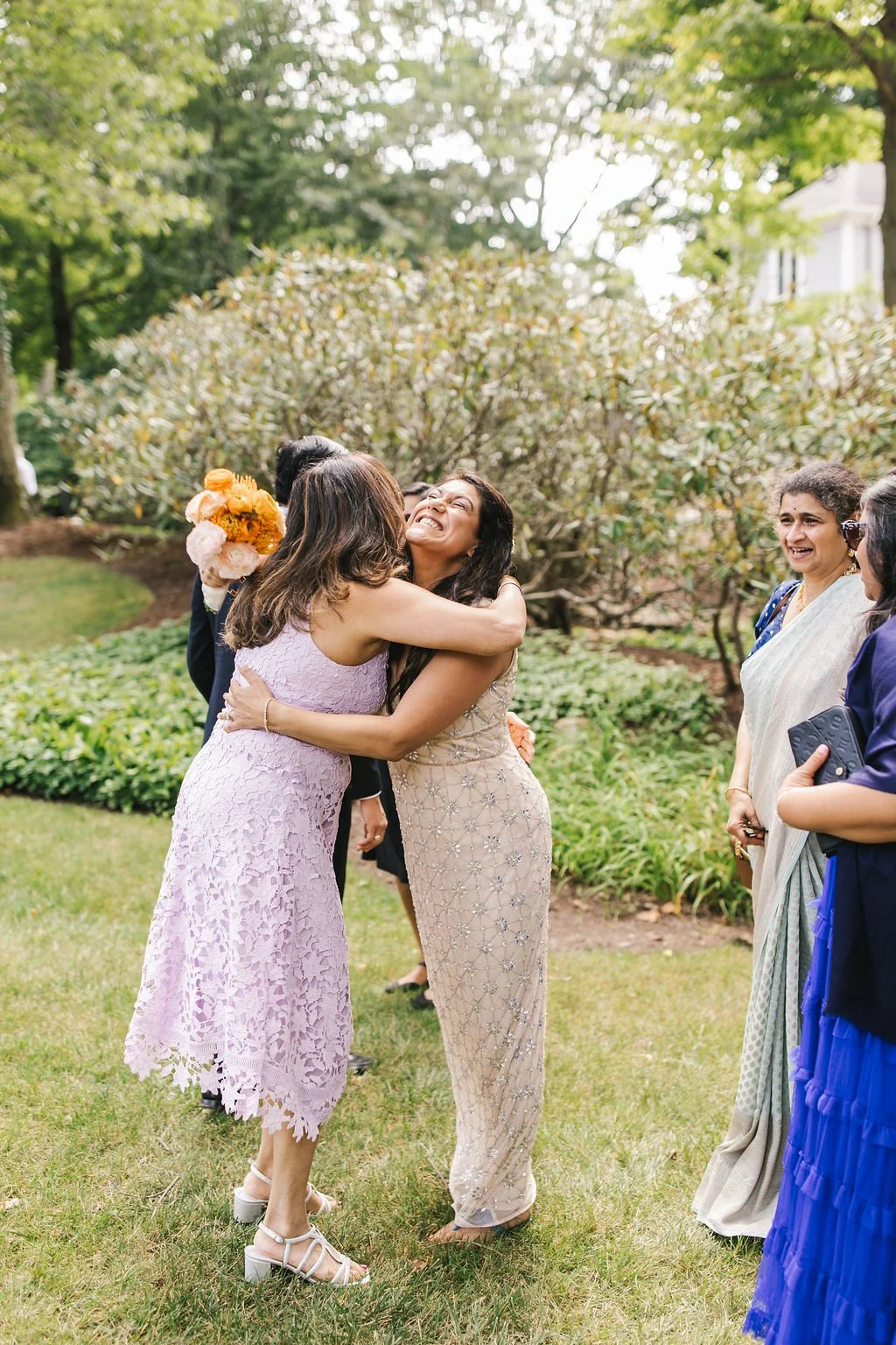 Two women embracing and smiling outdoors at a garden gathering, surrounded by other women and lush greenery.