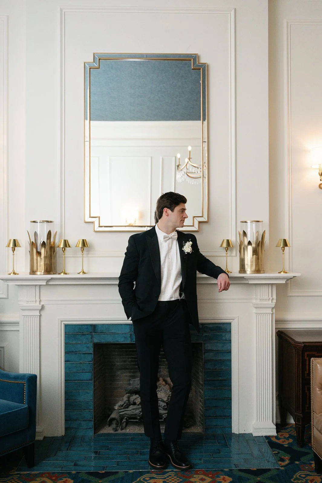 A man in a tuxedo stands in a decorated room by a fireplace, looking to his left with his hand resting on the mantel. The room features a large mirror above the fireplace, gold candlesticks, and gold vases on the mantel. There is a blue chair on the 