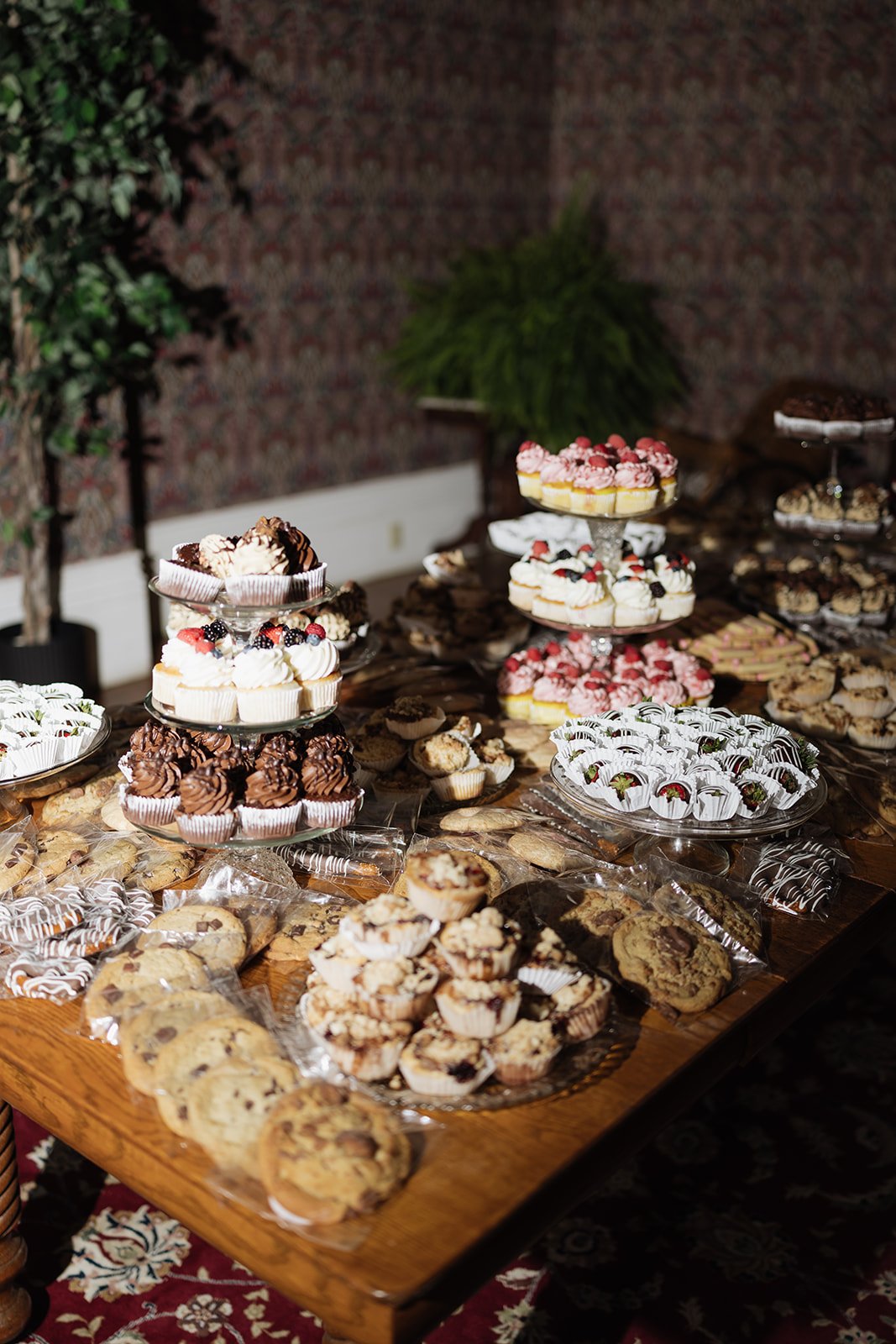 Assorted desserts on a wooden table, including cookies, cupcakes, and mini cakes on tiered glass stands at a party or celebration.