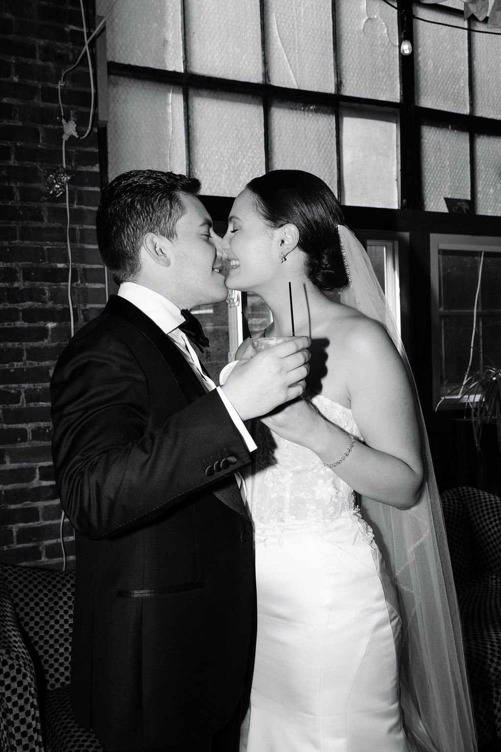 A black and white photo of a bride and groom sharing a kiss at their wedding reception. The groom is dressed in a tuxedo, and the bride is in a white wedding gown with a veil, holding a drink with two straws.