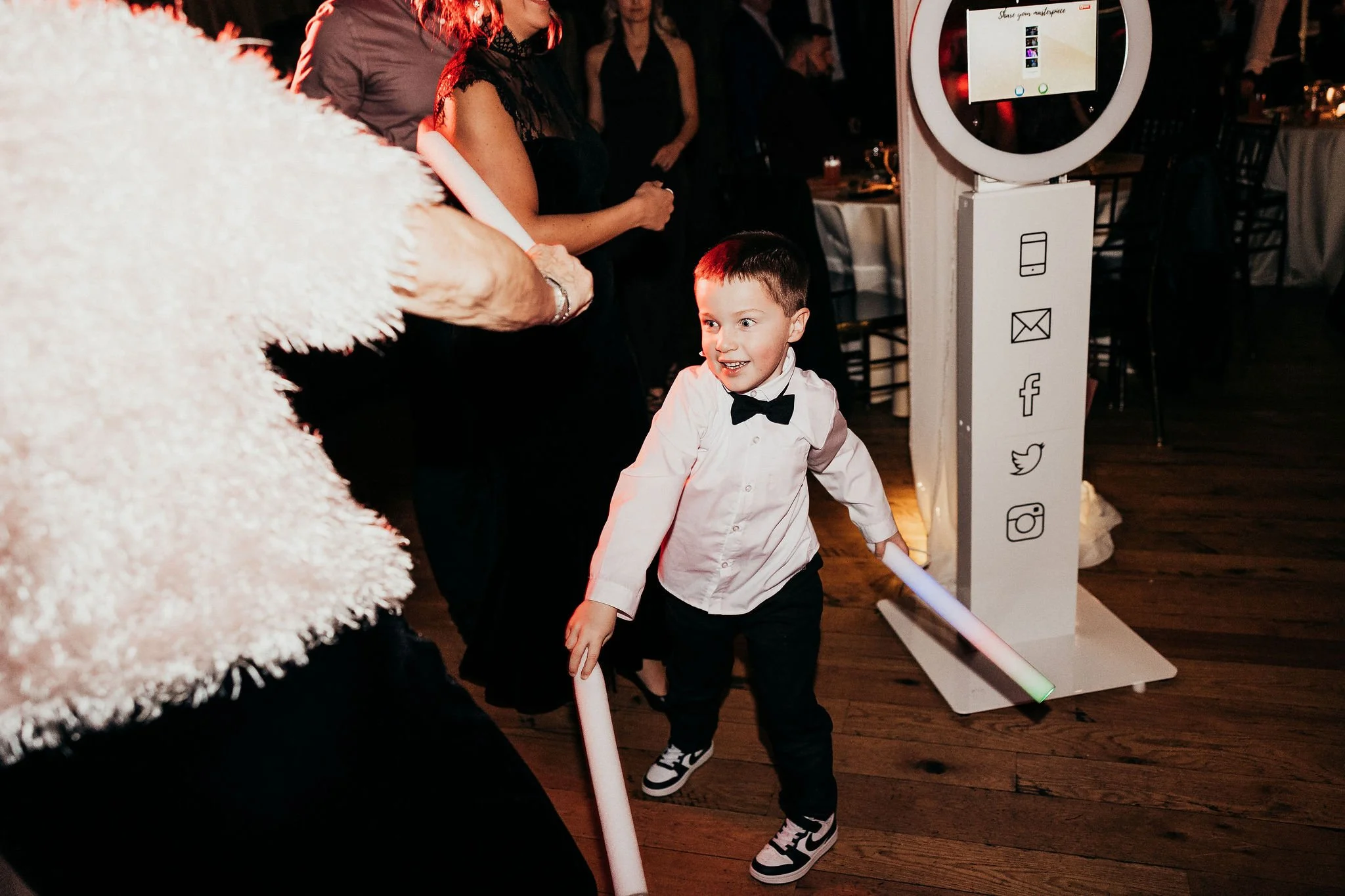 A young boy wearing a white shirt, black bow tie, and sneakers is dancing at a party, smiling and holding a light-up stick. He is surrounded by other people, including a woman in a black dress and a person dressed in a fluffy white costume. There is 