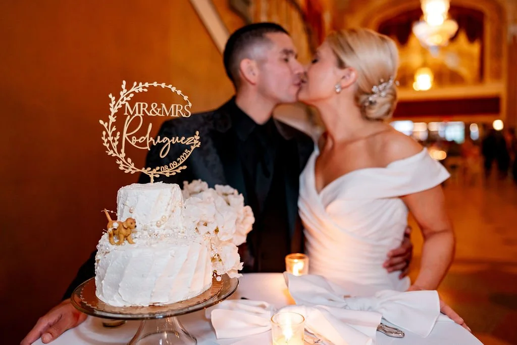 A newlywed couple kissing at their wedding reception with a wedding cake in the foreground, decorated with white flowers and a topper that reads 'Mr. & Mrs. Rodriguez 08.09.2023'.