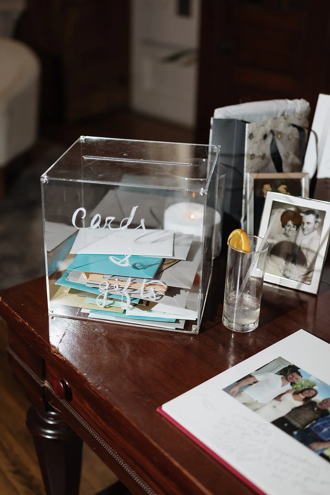 A transparent donation box labeled 'Cards & Gifts' with various envelopes inside, a photo frame with a wedding picture, a glass with a lemon wedge and straw, a wedding album, and gift bags on a wooden table.
