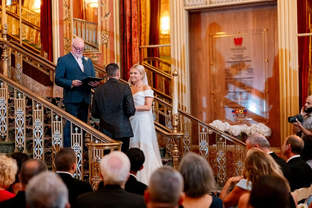 A wedding ceremony taking place indoors on a staircase with ornate golden railing, a man officiating, a woman in white wedding dress, and a man in a dark suit, with guests seated watching.
