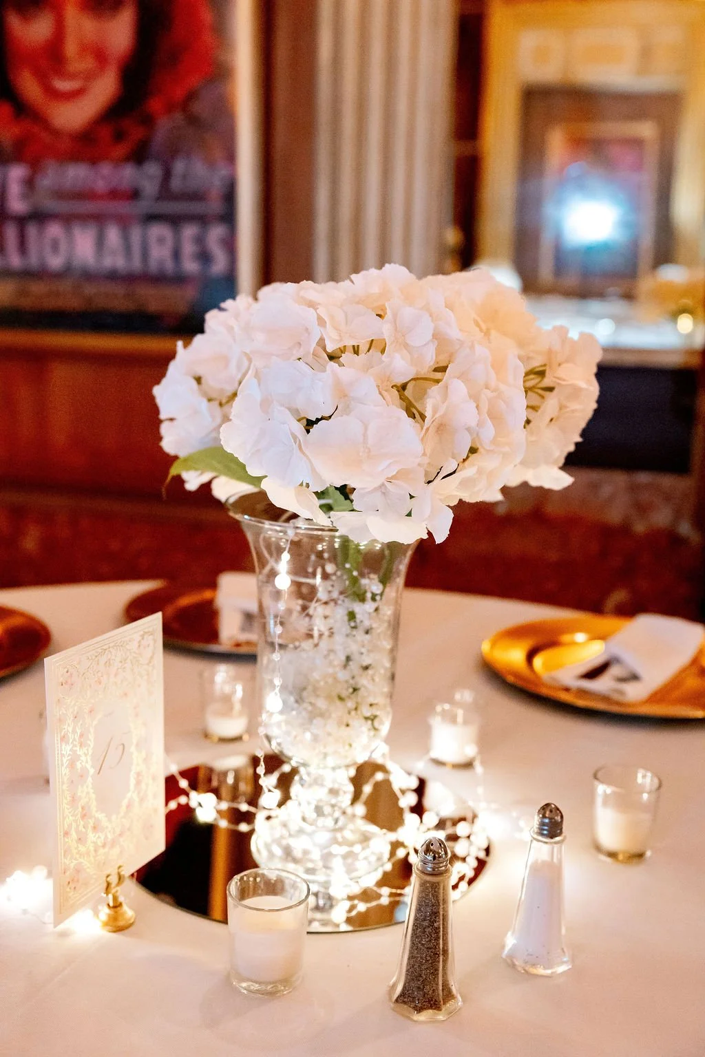 A round table decorated with a white hydrangea flower centerpiece in a tall glass vase, surrounded by small votive candles, salt and pepper shakers, gold plates, gold utensils, a folded napkin, and a table number card, with a mirror underneath the ce