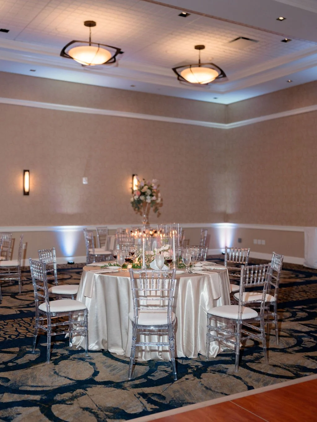 Elegant banquet table with white satin tablecloth, clear acrylic chairs, floral centerpiece, tableware, and wine glasses in a decorated banquet hall with soft lighting and floral decor