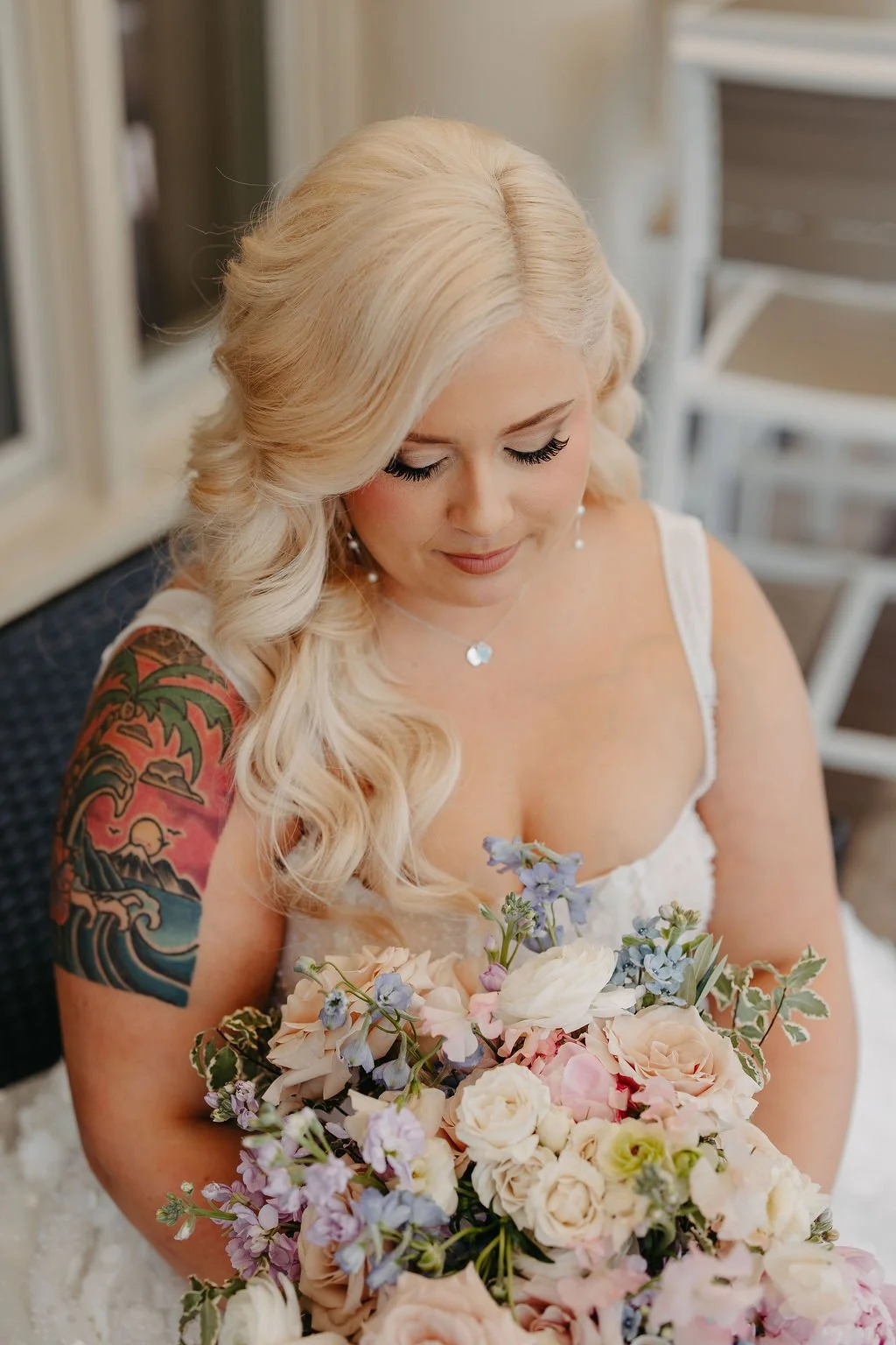 A blonde woman with wavy hair, wearing a white dress, holding a bouquet of pastel-colored flowers, and sitting on a chair.