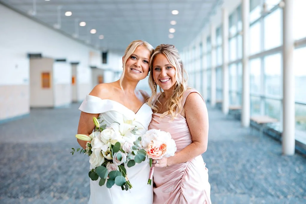 Two women smiling, one in a white wedding dress holding a bouquet, and the other in a light pink dress, standing in a modern, glass-walled corridor.