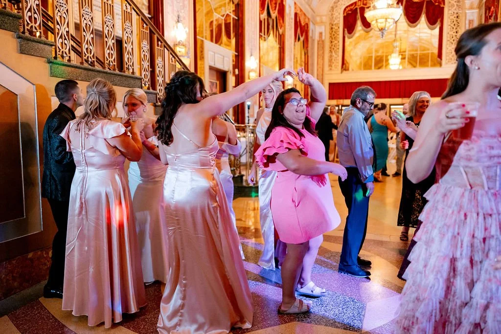 People dancing and socializing in a grand, ornate hall with chandeliers and intricate wall decorations during a celebration or wedding reception.