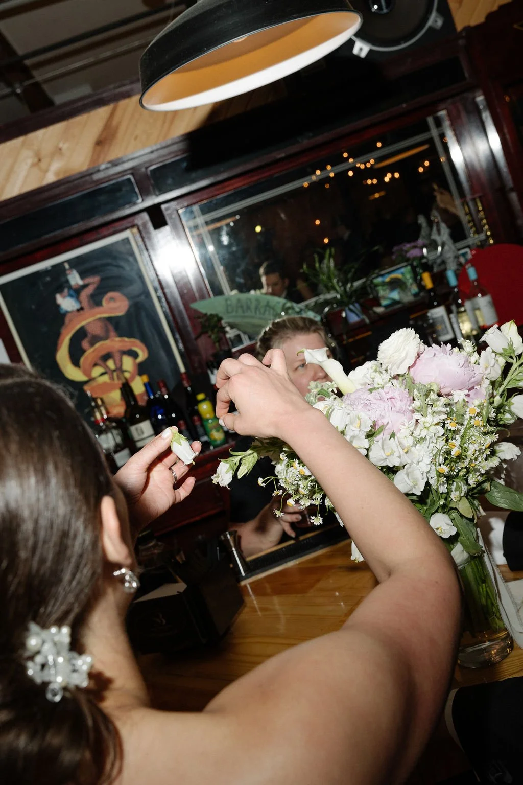 People arranging a flower bouquet at a bar counter in a restaurant or bar with artwork of a snake on the wall, liquor bottles on shelves, and string lights outside the window.